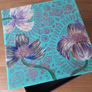 May include: A teal blue wooden box with a hinged lid. The lid is decorated with a hand-painted design of three purple flowers with white accents and a background of swirling patterns.