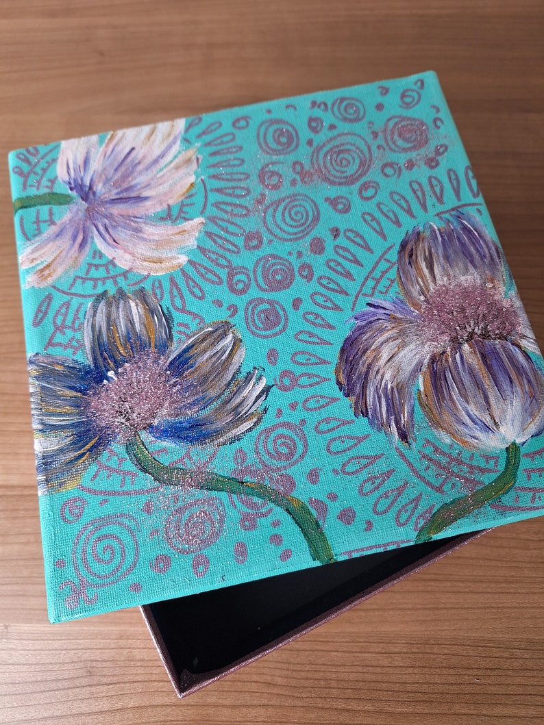 May include: A teal blue wooden box with a hinged lid. The lid is decorated with a hand-painted design of three purple flowers with white accents and a background of swirling patterns.