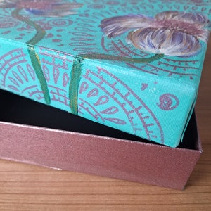 May include: A teal blue canvas box with a painted design of purple flowers and green leaves. The box is open and the lid is resting on top of a brown wooden surface.