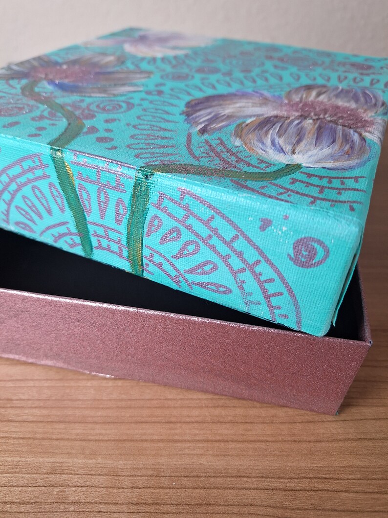 May include: A teal blue canvas box with a painted design of purple flowers and green leaves. The box is open and the lid is resting on top of a brown wooden surface.
