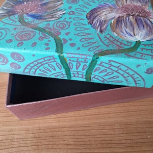 May include: Teal painted box with a floral design featuring two white flowers with purple centers. The box is open and the inside is brown.