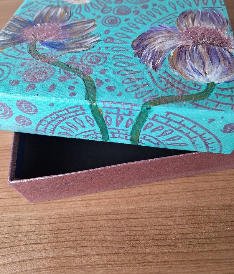 May include: Teal painted box with a floral design featuring two white flowers with purple centers. The box is open and the inside is brown.