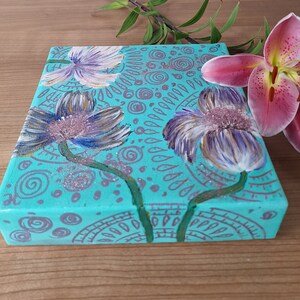 May include: A turquoise canvas painting with three stylized flowers in shades of purple, white, and pink. The background is decorated with a pattern of swirls and dots in a silver color.
