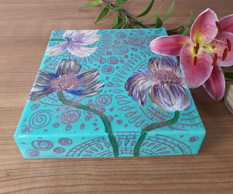 May include: A turquoise canvas painting with three stylized flowers in shades of purple, white, and pink. The background is decorated with a pattern of swirls and dots in a silver color.