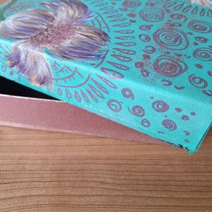 May include: A teal blue box with a purple flower painted on the lid. The box is decorated with a pattern of circles and lines in a darker shade of purple.