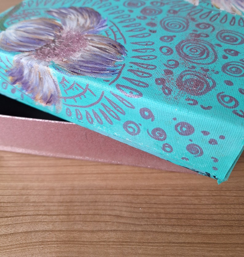 May include: A teal blue box with a purple flower painted on the lid. The box is decorated with a pattern of circles and lines in a darker shade of purple.
