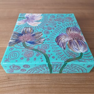 May include: A teal canvas painting with three purple and white flowers with green stems. The background is covered in a pattern of teal and purple circles and swirls.