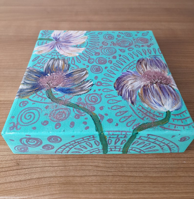 May include: A teal canvas painting with three purple and white flowers with green stems. The background is covered in a pattern of teal and purple circles and swirls.