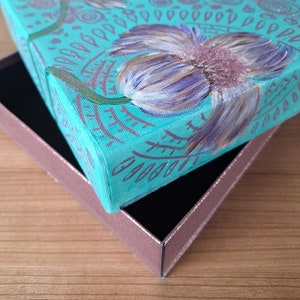 May include: A teal blue box with a painted design of a purple flower with green leaves. The box is open and the lid is resting on top of the box.