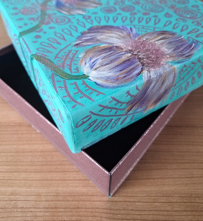 May include: A teal blue box with a painted design of a purple flower with green leaves. The box is open and the lid is resting on top of the box.