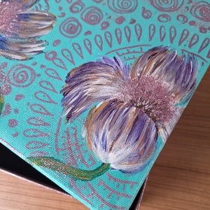 May include: A teal blue canvas box with a painted design of white and purple flowers with gold accents. The box has a glittery finish and is decorated with swirls and dots.