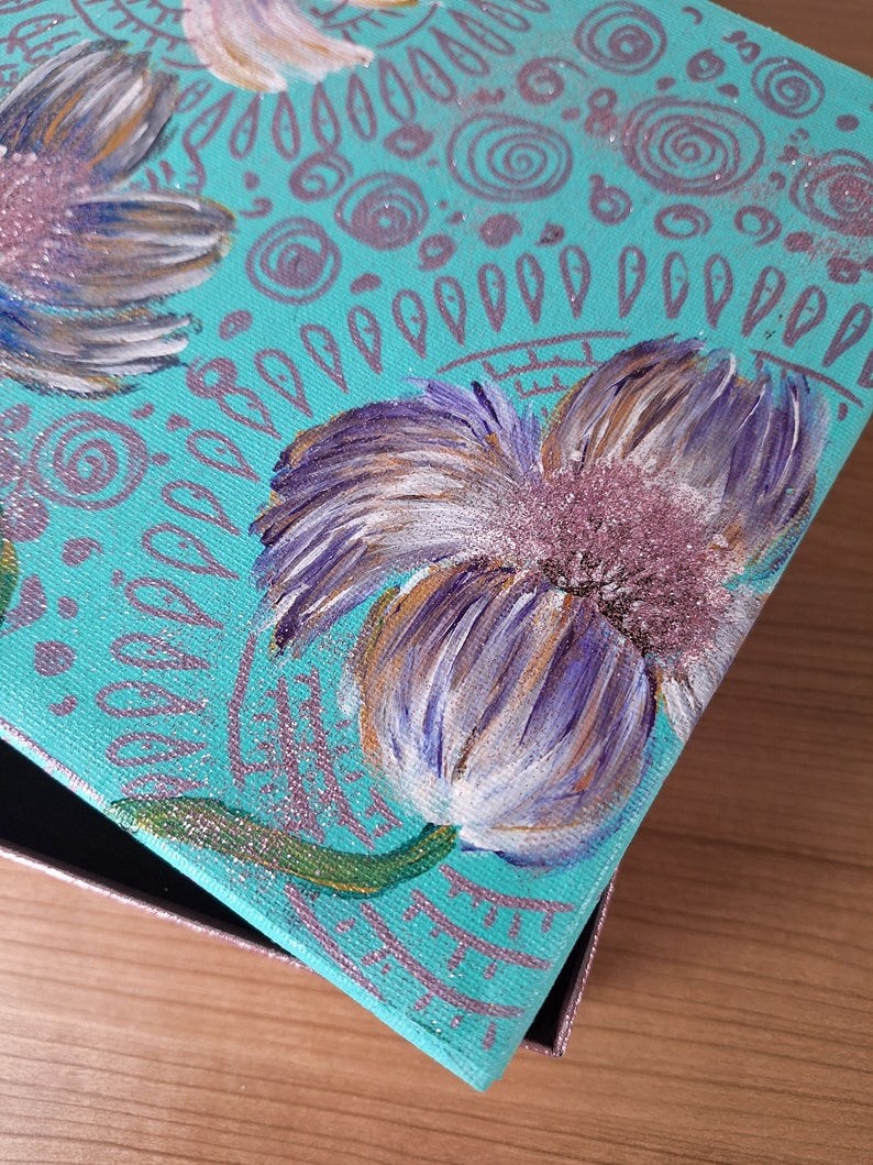 May include: A teal blue canvas box with a painted design of white and purple flowers with gold accents. The box has a glittery finish and is decorated with swirls and dots.
