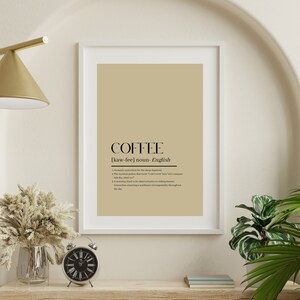 Könnte beinhalten: Ein beigefarbener gerahmter Druck mit dem Wort "COFFEE" in schwarzem Text. Die Definition von Kaffee ist darunter in schwarzem Text geschrieben. Der Druck hängt an einer weißen Wand mit einer goldenen Lampe, einer Vase mit Trockenblumen und einer Uhr auf einem Holzregal.