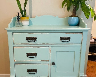May include: A light blue dresser with four drawers and a cabinet door. The dresser has distressed paint and black metal hardware. There are two potted plants on top of the dresser.