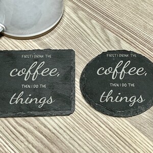 以下が含まれることがあります： 白い文字が書かれた黒いスレートコースター2枚。コースターの文字は「FIRST I DRINK THE coffee, THEN I DO THE things」です。
