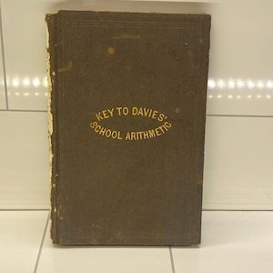 May include: An antique book with a worn, brown cover. The title "KEY TO DAVIES' SCHOOL ARITHMETIC" is printed in gold lettering. The book's edges show signs of age and wear, suggesting it is a vintage item.