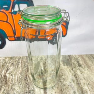 May include: A clear glass jar with a green-rimmed lid and a metal clasp. The jar has a cylindrical shape with vertical ridges. The lid is transparent, and the clasp is silver. The jar is empty and sits on a patterned surface.
