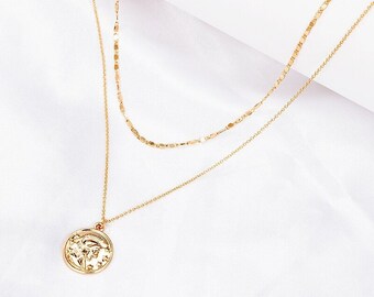 May include: A gold-toned layered necklace featuring two distinct chains. The shorter chain has a flat, textured link design, while the longer chain is a delicate cable style that holds a circular pendant with an embossed profile portrait.