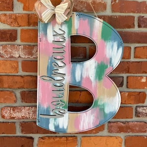 May include: A wooden letter B painted in shades of pink, blue, and green with a white background. The letter is decorated with a bow and the word "beautifully" is written on the side.
