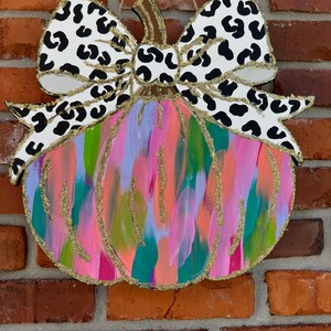 May include: A pink, green and blue abstract pumpkin with a black and white leopard print bow. The pumpkin is painted with a brushstroke effect and has gold glitter accents.