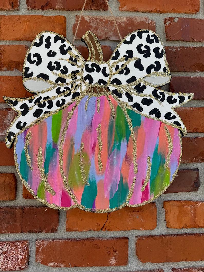 May include: A pink, green and blue abstract pumpkin with a black and white leopard print bow. The pumpkin is painted with a brushstroke effect and has gold glitter accents.
