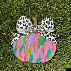 May include: A wooden pumpkin door hanger featuring a colourful abstract design and a black and white leopard print bow. The pumpkin is painted in shades of pink, green, blue, and orange with gold glitter accents.