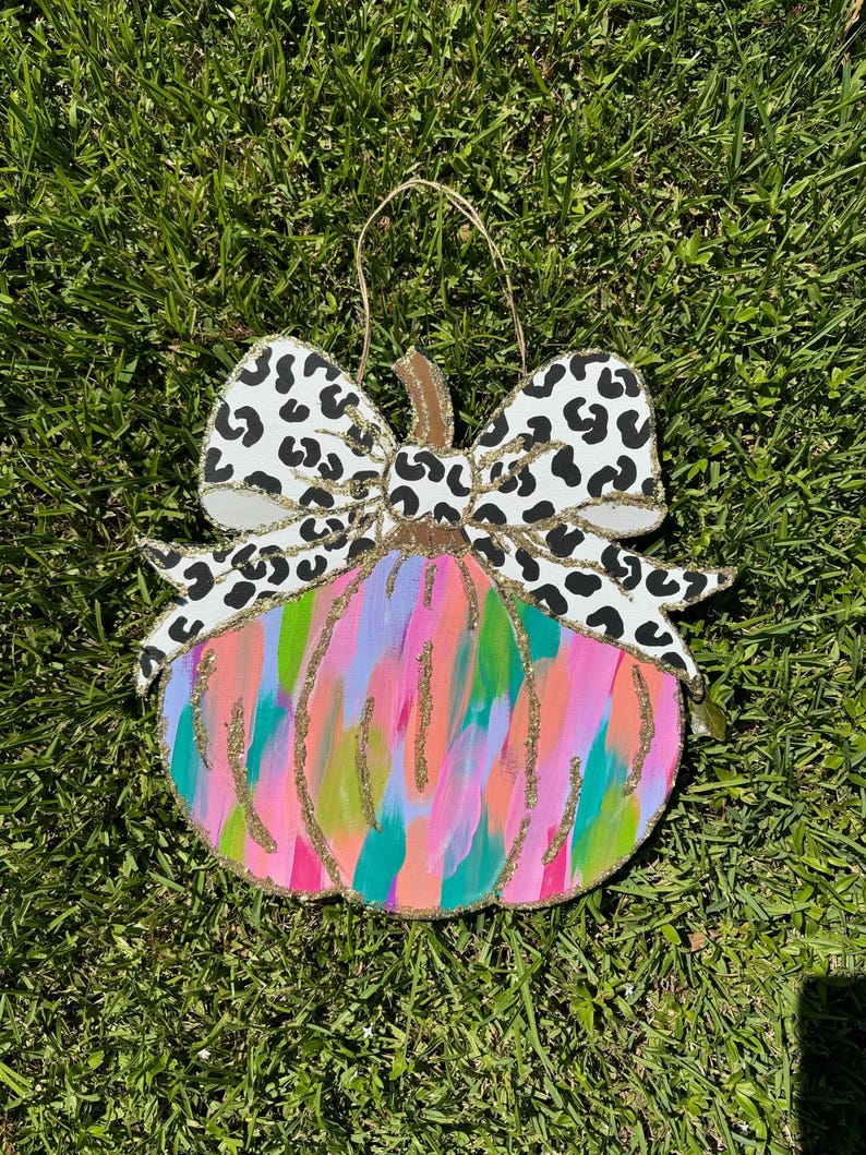 May include: A wooden pumpkin door hanger featuring a colourful abstract design and a black and white leopard print bow. The pumpkin is painted in shades of pink, green, blue, and orange with gold glitter accents.