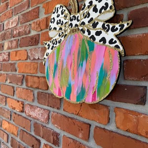 May include: A colourful, abstract pumpkin with a black and white leopard print bow. The pumpkin is painted with pink, blue, green, and gold paint.