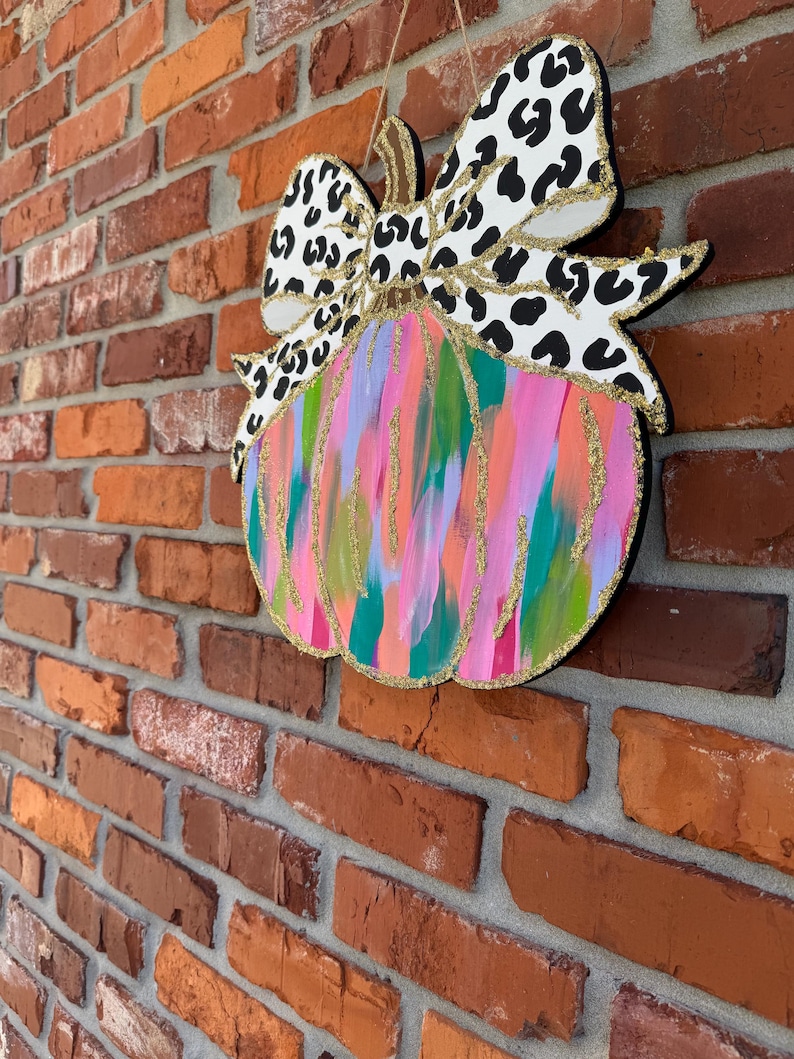 May include: A colourful, abstract pumpkin with a black and white leopard print bow. The pumpkin is painted with pink, blue, green, and gold paint.