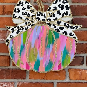 May include: A colourful, abstract pumpkin door hanger with a black and white leopard print bow. The pumpkin is painted in shades of pink, blue, green, and orange with gold glitter accents.