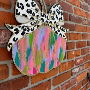 May include: A wooden pumpkin door hanger with a large black and white leopard print bow. The pumpkin is painted in shades of pink, green, blue, and gold with glitter accents.