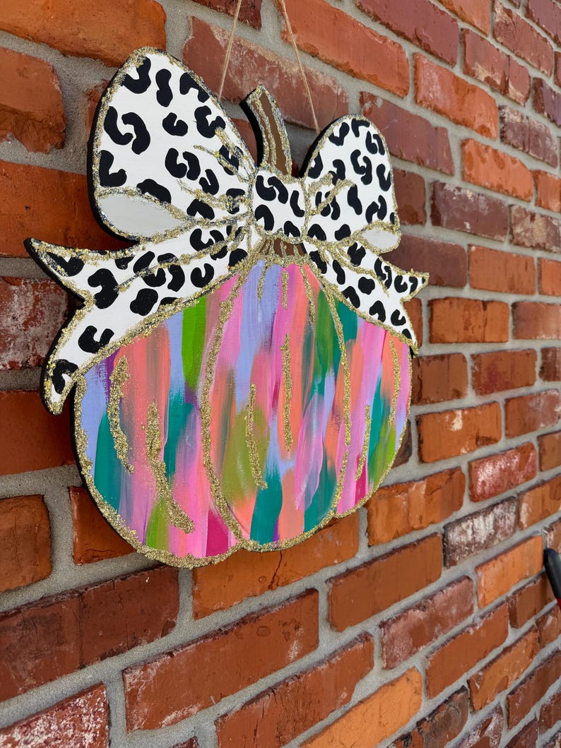 May include: A wooden pumpkin door hanger with a large black and white leopard print bow. The pumpkin is painted in shades of pink, green, blue, and gold with glitter accents.