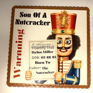 Puede incluir: Un letrero decorativo con un soldado cascanueces con un sombrero rojo y dorado y un uniforme azul. El letrero dice "Son Of A Nutcracker" e incluye un certificado con el nombre "Dylan Miller".