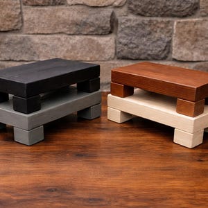 Op de afbeelding: Twee houten opstapjes. Eén is zwart op grijs, de andere bruin op crème. Elk krukje heeft een rechthoekige bovenkant die wordt ondersteund door vierkante poten. De krukjes worden op een houten oppervlak gepresenteerd.