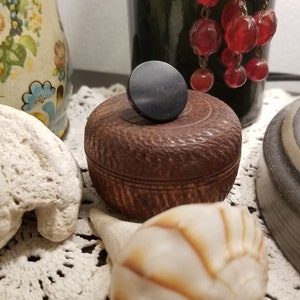 May include: A small, round, brown wooden box with a black knob on top. The box is intricately carved. A white seashell and a decorative pitcher are also visible.