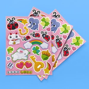 May include: A sheet of pink stickers featuring various illustrations including a ladybug, a white cat, a rabbit, a rainbow, a four-leaf clover, a yellow key, a die, and heart-shaped glasses. The text GOOD LUCK is printed at the bottom left.