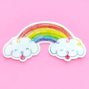 Könnte beinhalten: Ein farbenfroher Regenbogen- und Wolkenaufkleber auf rosa Hintergrund. Der Regenbogen zeigt rote, orange, gelbe, grüne, blaue und violette Farbtöne. Zwei weiße Wolken mit lächelnden Gesichtern und roten Zungen befinden sich am unteren Rand.