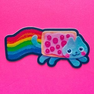 Puede incluir: Una pegatina caprichosa que presenta un gato azul con un cuerpo de Pop-Tart y una cola de arcoíris. El Pop-Tart tiene glaseado rosa y cobertura de bayas rojas. El gato tiene una cara sonriente con mejillas rosadas y la pegatina está delineada en negro.