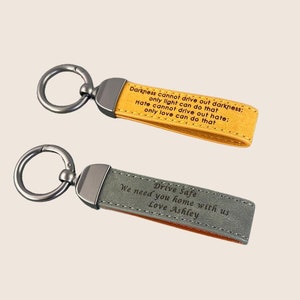 Puede incluir: Dos llaveros de cuero con anillos de metal. El llavero superior es amarillo con el texto "Darkness cannot drive out darkness; only light can do that. Hate cannot drive out hate; only love can do that." El llavero inferior es gris con el texto "Drive Safe. We need you home with us. Love Ashley."
