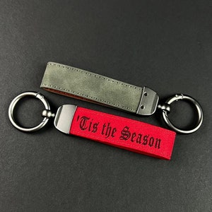 Könnte beinhalten: Zwei Schlüsselanhänger mit Metallringen und Lederriemen. Ein Riemen ist rot mit dem Text "'Tis the Season" in einer dekorativen Schriftart. Der andere Riemen ist olivgrün. Beide haben silberfarbene Metallbeschläge.