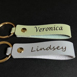 May include: Two personalized keychains with gold-colored rings. One is light green with the name "Veronica" and the other is light gray with the name "Lindsey". Both keychains have a gold-colored snap.
