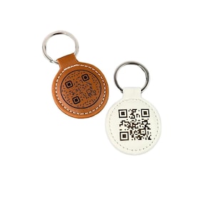 Puede incluir: Dos llaveros de cuero con forma redonda y un anillo de metal. Un llavero es marrón con un código QR negro y el otro es blanco con un código QR negro.