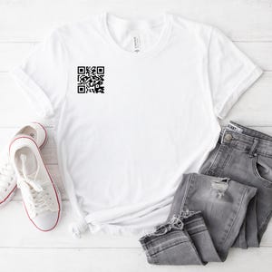 May include: A white t-shirt with a black QR code printed on the front. The shirt is lying on a white wooden surface with a pair of white trainers and a pair of grey jeans.