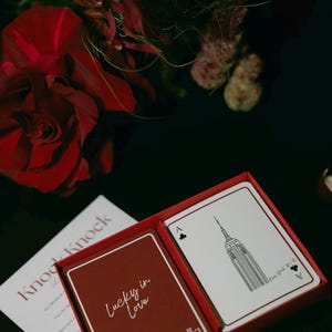 May include: A red playing card box with two decks of cards. One deck has the words "Lucky in Love" on a red background. The other deck has a black and white illustration of the Empire State Building. A red rose and floral arrangement are in the background.