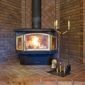 May include: A black and gold wood-burning stove with a glass door, set within a brick fireplace. The fire is lit, with flames visible inside. A set of fireplace tools, including a poker, shovel, and brush, sits on a black metal stand in front of the stove.