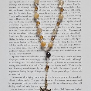 May include: A necklace with a sun pendant and various beads. The pendant is a detailed, antique gold-colored sunburst design. The beads are a mix of light brown, clear, and black, strung on a delicate chain. The necklace is displayed on a page of text.