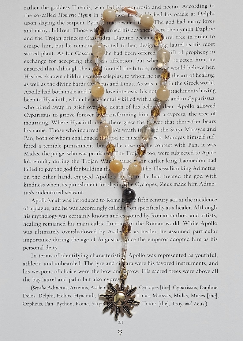 May include: A necklace with a sun pendant and various beads. The pendant is a detailed, antique gold-colored sunburst design. The beads are a mix of light brown, clear, and black, strung on a delicate chain. The necklace is displayed on a page of text.