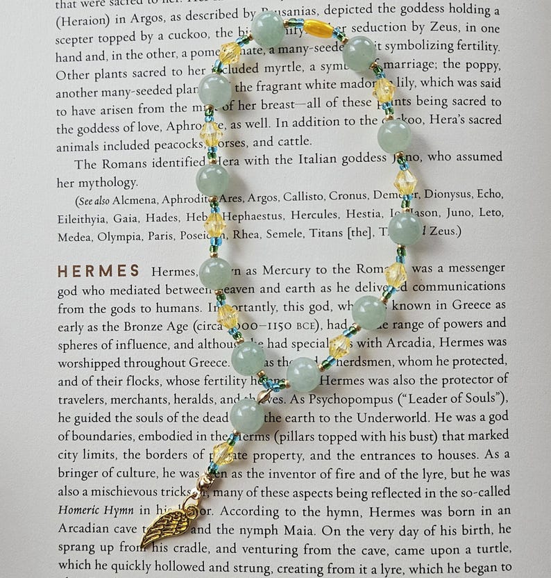 May include: A beaded necklace featuring light green and yellow beads. The necklace has a gold-colored charm with a wing design. The necklace is displayed on top of a book.
