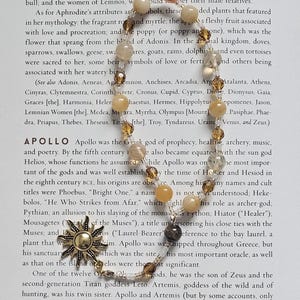 May include: A beaded necklace with a gold-tone sun pendant and various light-colored beads. The necklace is displayed on a page of text, likely from a book, with the word "Apollo" visible.