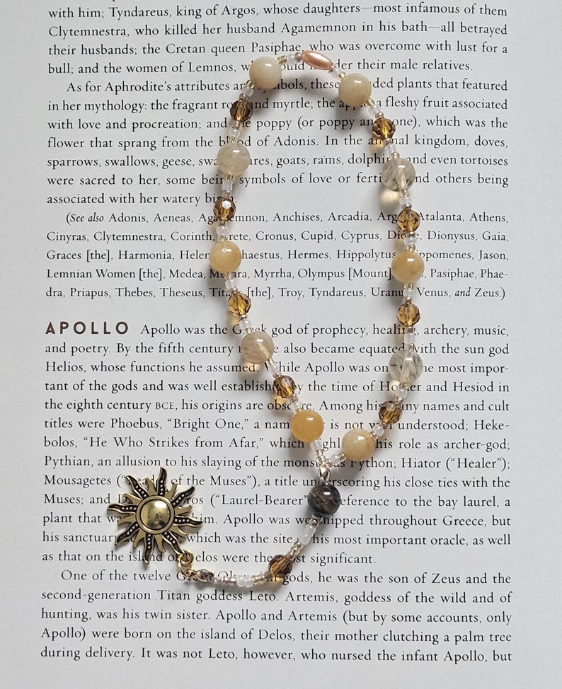 May include: A beaded necklace with a gold-tone sun pendant and various light-colored beads. The necklace is displayed on a page of text, likely from a book, with the word "Apollo" visible.